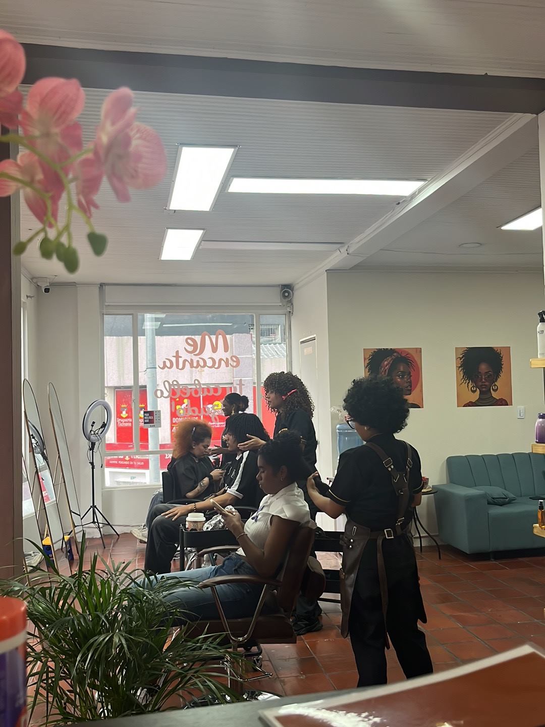 Interior del salón: estilistas trabajando con varias clientas, sofá verde y cuadros de mujeres afro